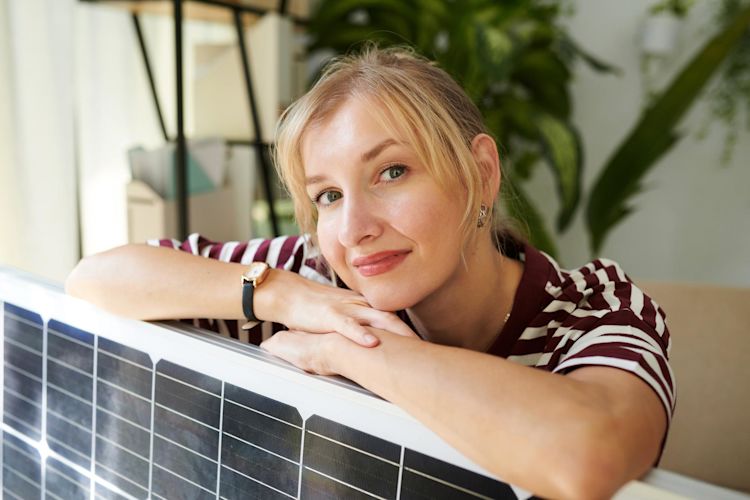 Grüner Strom mit Balkonsolaranlage kann so easy sein. Und Klimaschutz auch Junge Frau mit blonden Haaren lehnt zufrieden über ihrem Solarpanel in ihrer Wohnung
