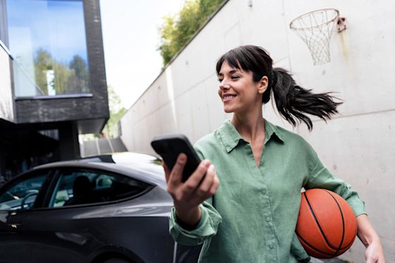 Strom von heute geht so: Mit flexiblen Tarifen und Smart Meter lädst du dein E-Auto günstiger Lachende Frau mit Basketball unter dem Arm und Handy in der Hand lädt ihr E-Auto smart zuhause